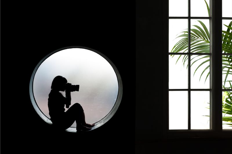 A silhouette of a person holding a camera, sitting in a round window, with a bright window and plant visible in the background.