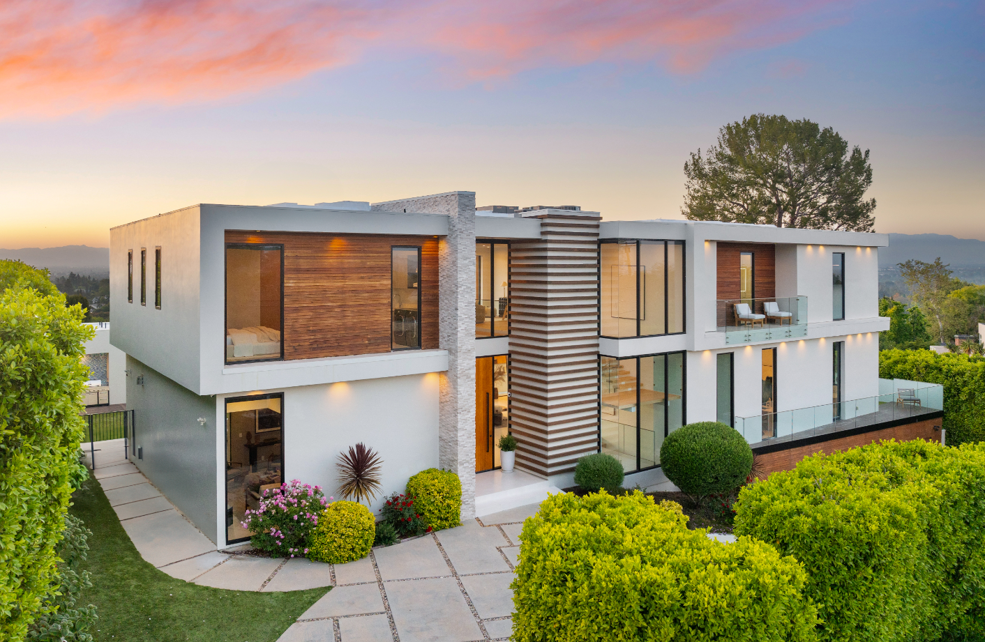 A stylish, modern two-story home with glass panels, wooden accents, and lush landscaping, captured at sunset with a serene sky in the background.