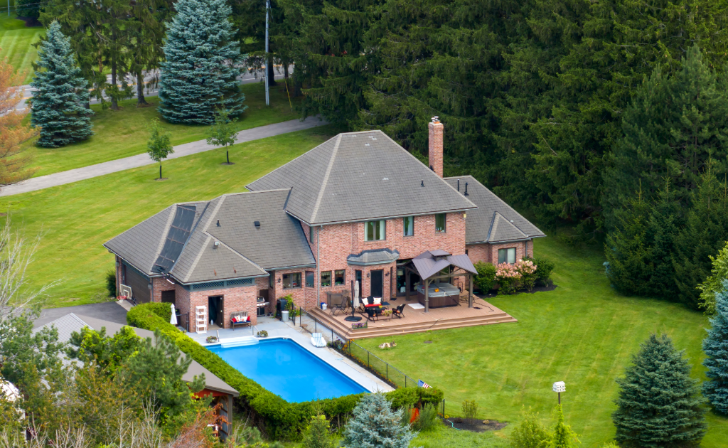 A large brick suburban house with a well-maintained lawn, a backyard patio, and a rectangular swimming pool surrounded by trees.